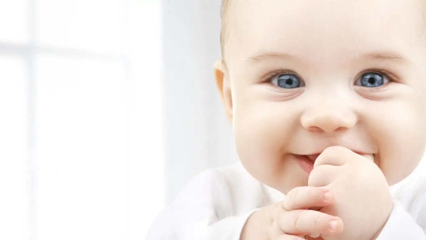 Щастливо бебе с големи сини очи и бяла блуза, което си смуче пръста. Консултации за плавни преходи в режима на сън и будуване през първата година.