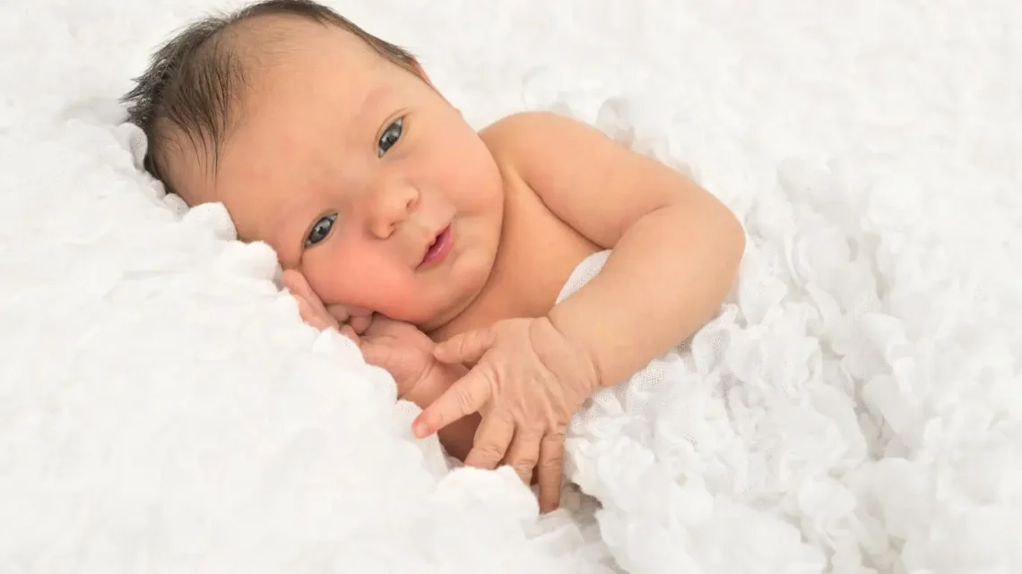 Усмихнато бебе, лежащо на бяла пухкава постелка. Консултации за самоуспокояване и самостоятелно заспиване, за да се подобри сънят на бебетата.